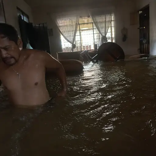 Sala inundada de agua oscura con muebles sumergidos y una persona en pleno caos.