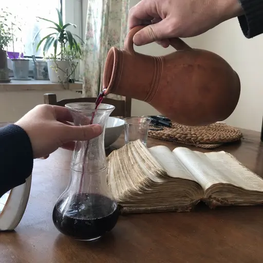Manos trasvasando vino de una jarra rústica junto a una Biblia antigua abierta.